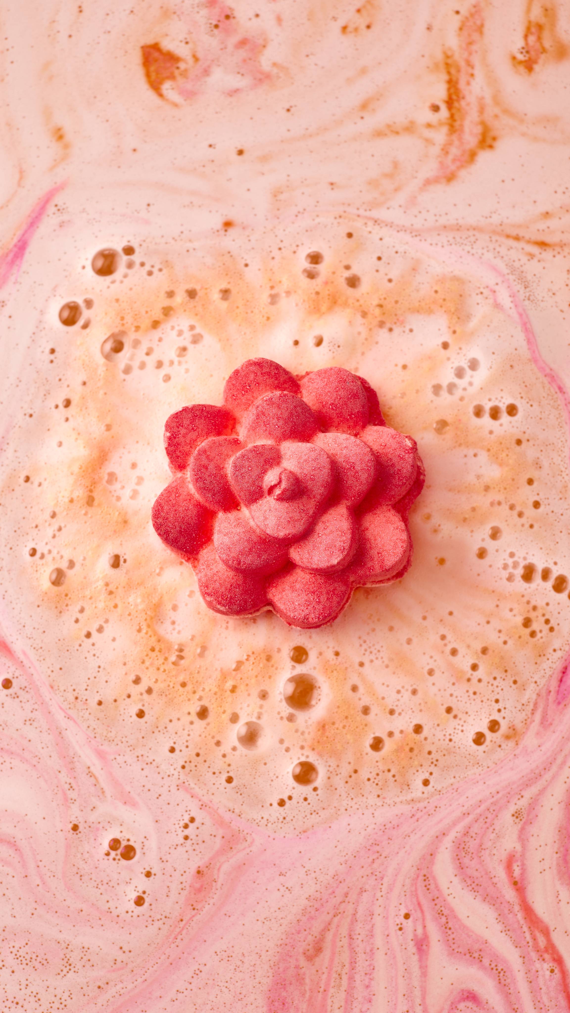 An aerial image of the "Comforter Rose' bath bomb as it begins to dissolve. It is surrounded by a ring of thick, cream-coloured bubbles, with swirling patterns of soft pink and peach radiating across the water's surface.