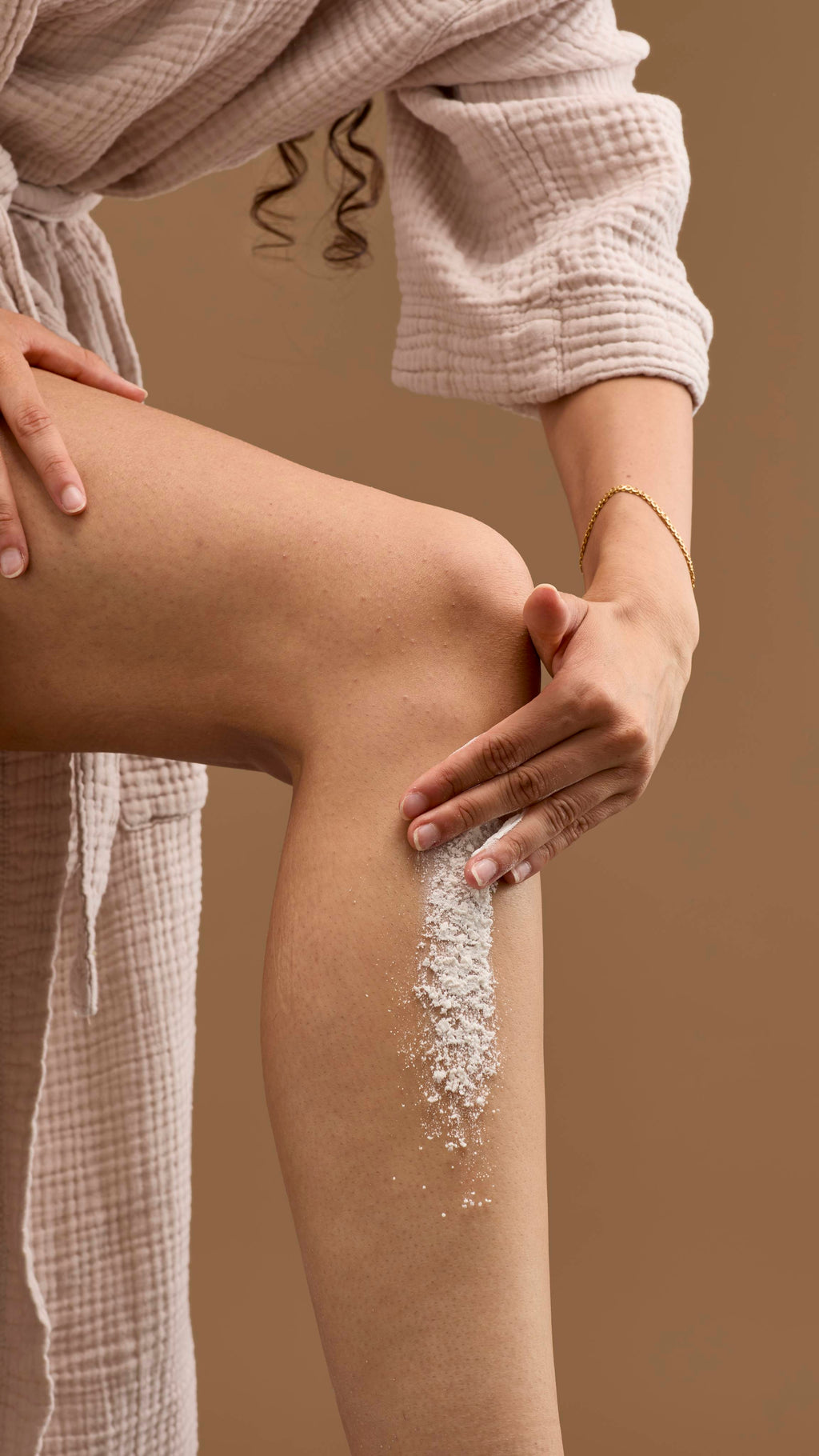 The image shows a hand applying a thick streak of creamy 'Silky Underwear' dusting powder onto a bare leg. The person wears a light-coloured robe and a bracelet against a solid beige background.