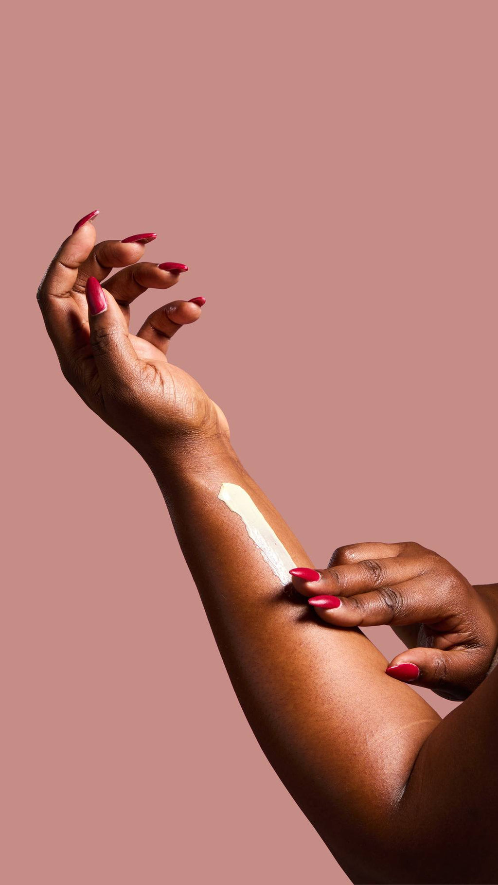 A hand with long, deep red nails massages a streak of creamy, sticky dates lotion into the skin of a forearm against a dusty pink background.