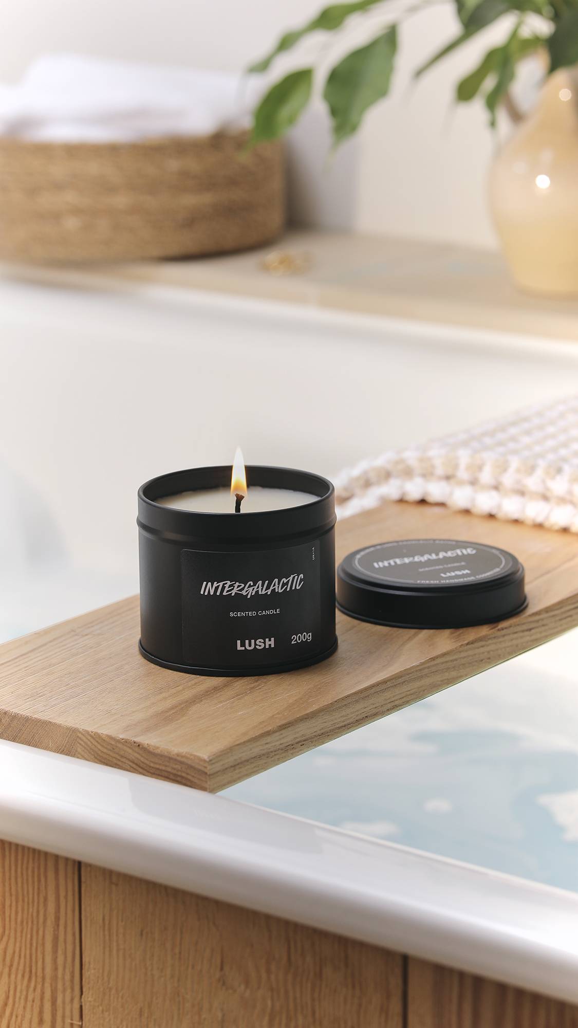 The image shows a bright, blurred bathroom scene with an out-of-focus wicker basket and plant. The black tin Intergalactic candle has a lit wick and is sitting on a wooden bath caddy laid over a filled bath.